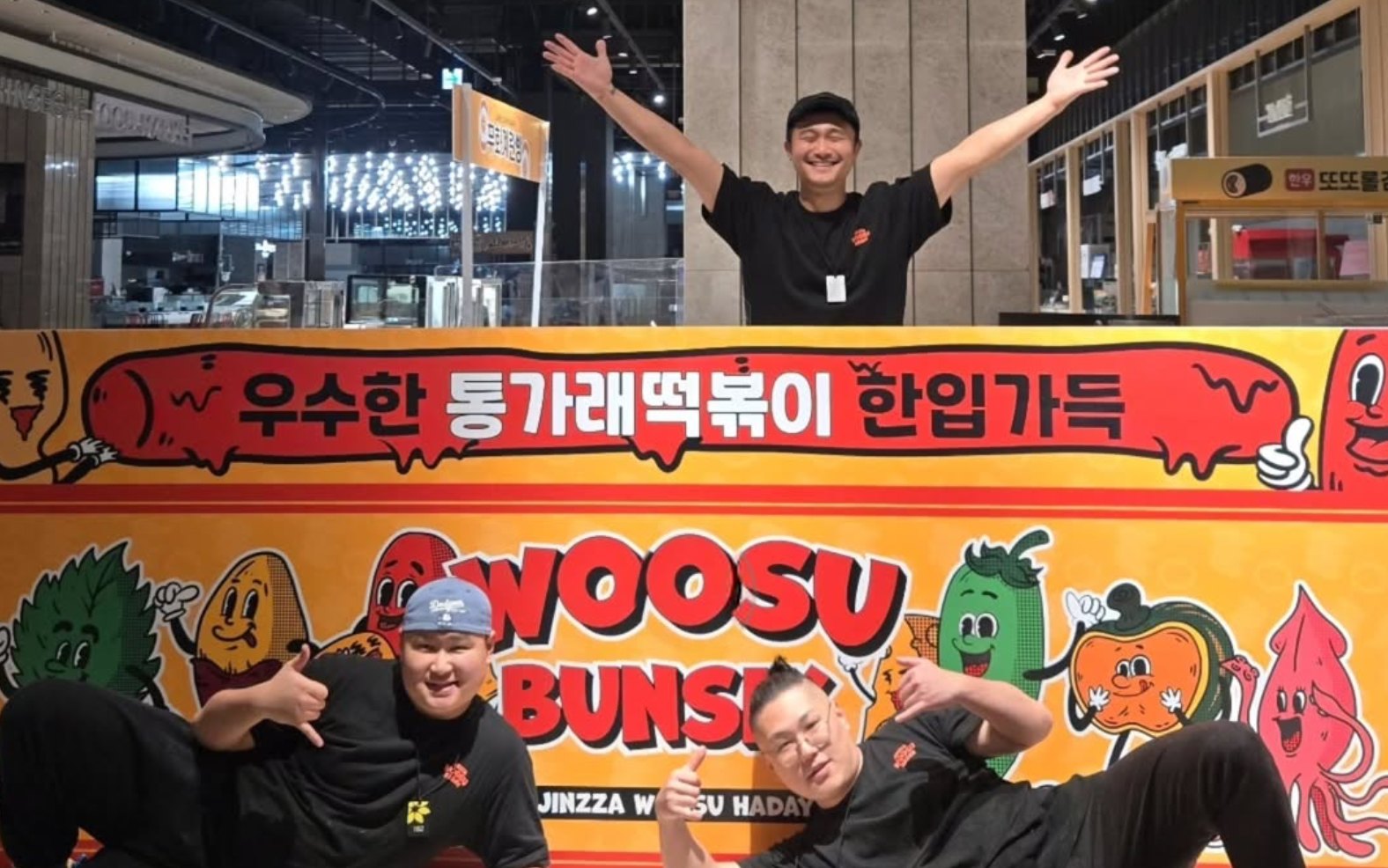 대구 신세계 팝업스토어의 WOOSU BUNSIK 벽화 앞에서 두 팔을 벌린 양윤석 대표와 바닥에 누워 포즈를 취한 팀원 두 명
